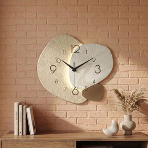 Può includere: Orologio da parete astratto con quadrante color crema e bianco, lancette nere e design minimalista. L'orologio ha un aspetto strutturato e stratificato ed è montato su un muro di mattoni. Il quadrante dell'orologio ha i numeri da 1 a 12.