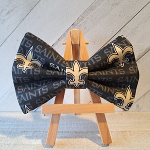 May include: A black bow tie with a gold fleur-de-lis and the word "Saints" repeated on the fabric. The bow tie is displayed on a wooden easel.