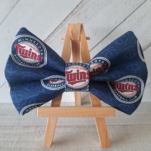 May include: A blue bow tie with a white and red Minnesota Twins baseball logo. The bow tie is on a wooden easel.