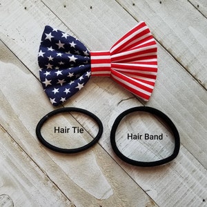 May include: A red, white, and blue bow tie with stars and stripes. The bow tie is on a white wooden surface with two black hair ties, one labeled "Hair Tie" and the other labeled "Hair Band".
