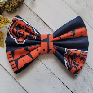 May include: A navy blue fabric bow tie featuring an orange bear head logo and the orange text "BEARS" printed across the surface. The bow tie has a central fabric knot and a repeating pattern of the team logo and text throughout the design.