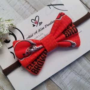 May include: A brown and orange bow with a football helmet and the words "Cleveland Browns" printed on it. The bow is attached to a brown elastic headband.