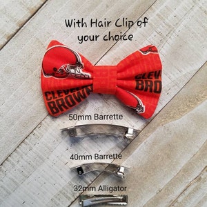 May include: Orange and brown bow tie hair clip with the text "CLEVELAND BROWN" and a football helmet design. The clip is available in three sizes: 50mm barrette, 40mm barrette, and 32mm alligator clip.