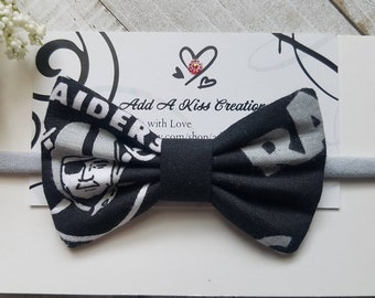 May include: Black and white bow tie headband with a Raiders logo. The headband is made of fabric and has a black elastic band.