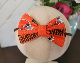 May include: Orange and brown bow with a Cleveland Browns logo and the words "Cleveland Browns" printed on it. The bow is on a wooden stand.