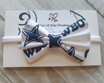 May include: White headband with a blue and white bow featuring the Dallas Cowboys logo. The bow is made of fabric with the Cowboys star and "Cowboys" text.