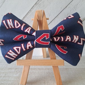 May include: A blue bow tie with a white and red "Indians" logo. The bow tie is displayed on a wooden easel.