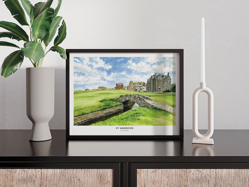May include: A framed watercolor print of St Andrews golf course, featuring a stone bridge over a small stream, green grass, and buildings under a blue sky. The print is in a black frame and is displayed on a dark surface.