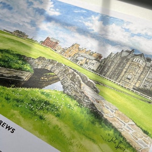 May include: Watercolor print of St Andrews, Scotland, featuring a stone bridge over a small stream, lush green grass, and historic buildings. The sky is blue with scattered clouds. The print includes the text "ST ANDREWS SCOTLAND".