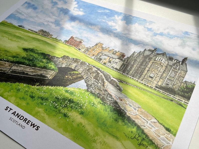 May include: Watercolor print of St Andrews, Scotland, featuring a stone bridge over a small stream, lush green grass, and historic buildings. The sky is blue with scattered clouds. The print includes the text "ST ANDREWS SCOTLAND".