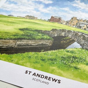 May include: Watercolor print of St Andrews, Scotland, featuring a stone bridge over a small stream, lush green grass, and buildings in the background. The sky is blue with scattered clouds. The text "ST ANDREWS SCOTLAND" is printed at the bottom.