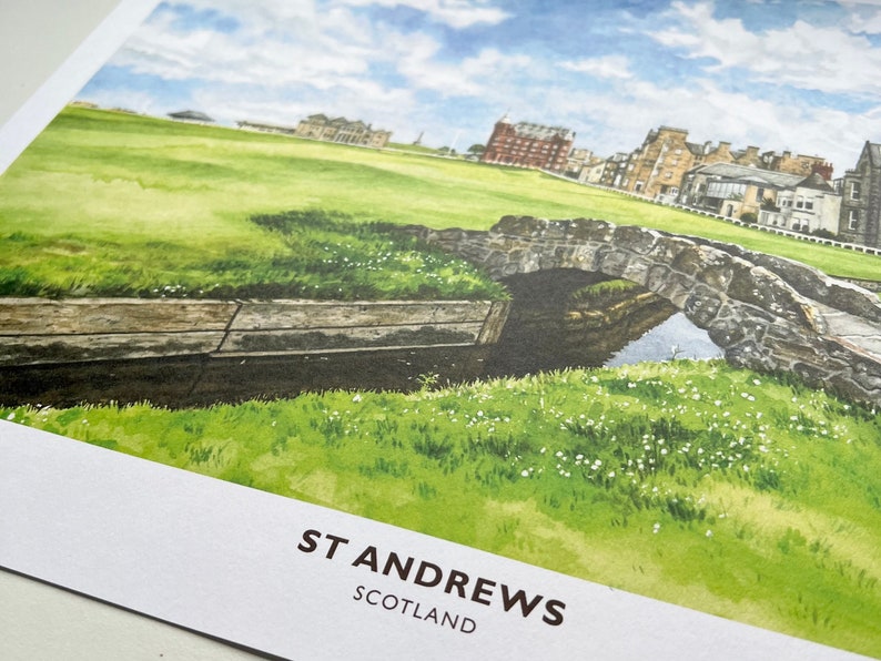 May include: Watercolor print of St Andrews, Scotland, featuring a stone bridge over a small stream, lush green grass, and buildings in the background. The sky is blue with scattered clouds. The text "ST ANDREWS SCOTLAND" is printed at the bottom.