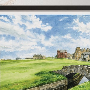 May include: Watercolor painting of a golf course with a stone bridge over a small stream in the foreground. Buildings and a cloudy blue sky are in the background. The artwork is framed in a black frame.