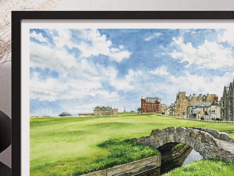 May include: Watercolor painting of a golf course with a stone bridge over a small stream in the foreground. Buildings and a cloudy blue sky are in the background. The artwork is framed in a black frame.