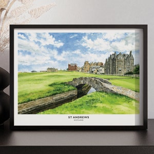 May include: A framed watercolor print of St Andrews, Scotland, featuring a stone bridge over a small stream, lush green golf course, and historic buildings under a blue sky with clouds. The print is in a black frame.