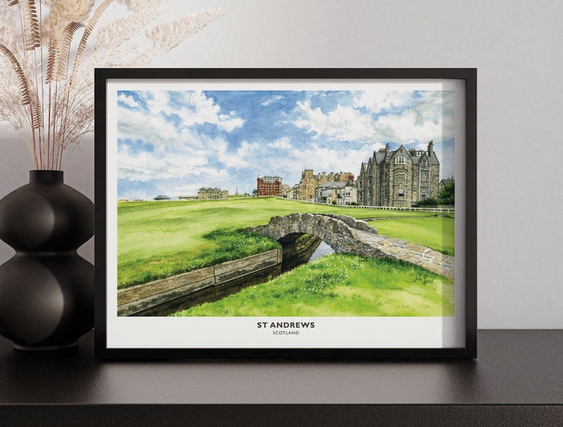 May include: A framed watercolor print of St Andrews, Scotland, featuring a stone bridge over a small stream, lush green golf course, and historic buildings under a blue sky with clouds. The print is in a black frame.