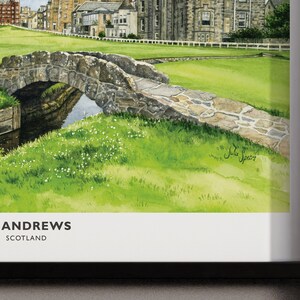 May include: Watercolor painting of St Andrews, Scotland, featuring a stone bridge over a small stream, lush green grass, and historic buildings. The artwork is framed in a black frame. The text "ST ANDREWS SCOTLAND" is at the bottom.