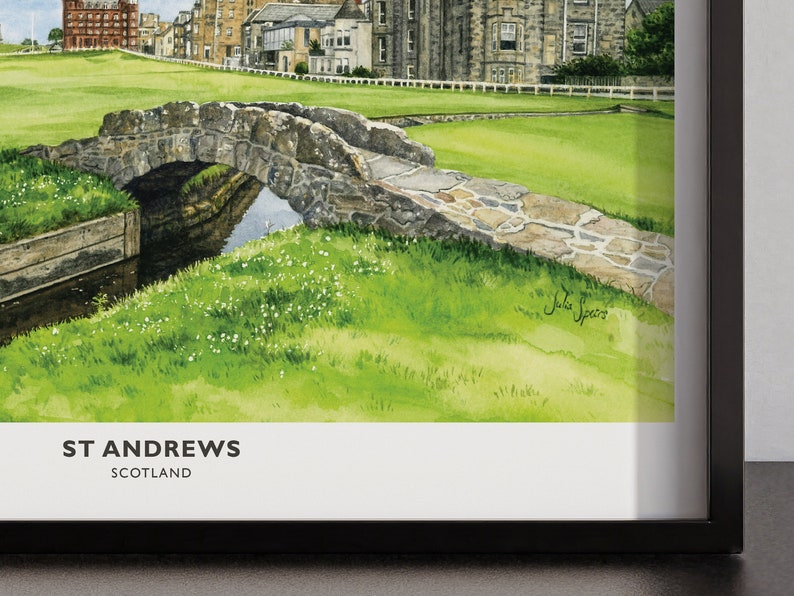 May include: Watercolor painting of St Andrews, Scotland, featuring a stone bridge over a small stream, lush green grass, and historic buildings. The artwork is framed in a black frame. The text "ST ANDREWS SCOTLAND" is at the bottom.