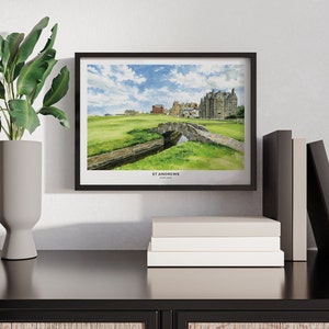 May include: A framed watercolor print of the St Andrews golf course, featuring a stone bridge over a small stream, green grass, and a cloudy blue sky. The print is in a black frame and is displayed on a black surface with books and a decorative plant.