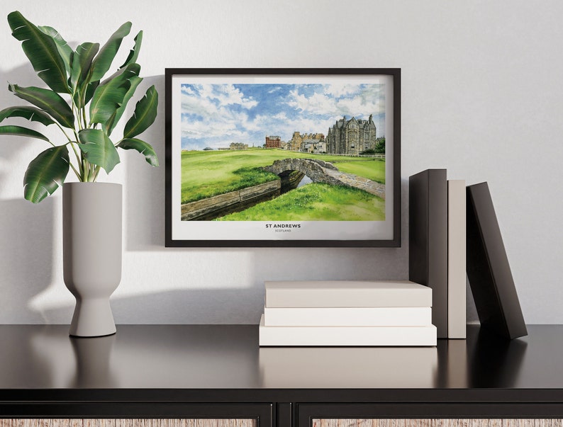 May include: A framed watercolor print of the St Andrews golf course, featuring a stone bridge over a small stream, green grass, and a cloudy blue sky. The print is in a black frame and is displayed on a black surface with books and a decorative plant.