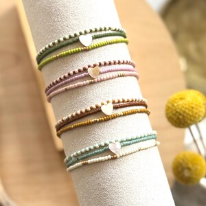 Può includere: Una pila di braccialetti di perline nei toni del verde, rosa, marrone e blu. Ogni braccialetto presenta un piccolo ciondolo a forma di cuore. I braccialetti sono esposti su un supporto cilindrico bianco, con sfondo sfocato.