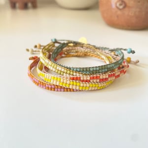 Könnte beinhalten: Ein Stapel bunter Perlenarmbänder in den Farben Gelb, Orange, Rosa, Grün und Blau. Die Armbänder sind aus kleinen, quadratischen Perlen gefertigt und übereinander gelegt.