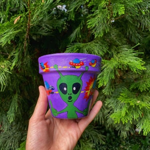 Puede incluir: Maceta de cerámica morada pintada a mano con un diseño de alienígena verde. La maceta presenta ilustraciones coloridas de platillos volantes y explosiones. Un objeto decorativo original para los amantes de las plantas.