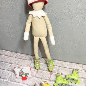 May include: A doll elf wearing a red hat with the name "Tabitha" embroidered on it, standing on green and yellow roller skates. The elf is holding a sign that says "Live Love Skate".