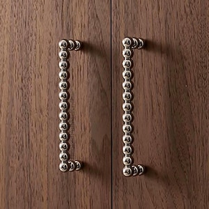 Pode incluir: Close-up de duas portas de armário de madeira castanha escura com puxadores verticais prateados com contas. Os puxadores são uniformemente espaçados e adicionam um toque de elegância.