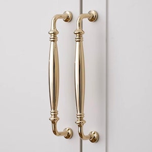 May include: Two gold cabinet pulls with a simple, elegant design. The pulls have a rounded, slightly curved shape and a smooth, polished finish.