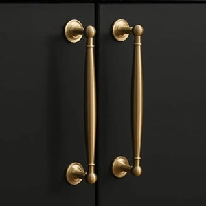 Op de afbeelding: Twee messing kastgrepen met een klassiek ontwerp. Elke greep heeft een lange, slanke handgreep met afgeronde uiteinden en decoratieve cirkelvormige achterplaten. De grepen zijn gemonteerd op een zwarte kastdeur.