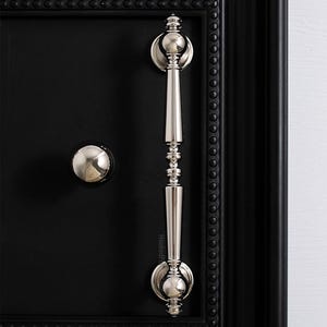 Pode incluir: Close-up de uma porta preta com puxador e maçaneta prateados. A maçaneta é um puxador vertical longo com detalhes decorativos. A porta é emoldurada com uma borda preta com um design de contas.