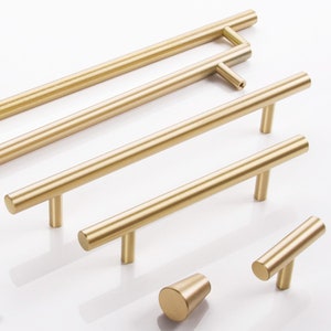 Puede incluir: Un juego de cinco tiradores de armario de color dorado en varios tamaños. Los tiradores están hechos de metal y tienen un acabado cepillado. Los tiradores están dispuestos en un patrón escalonado sobre una superficie blanca.