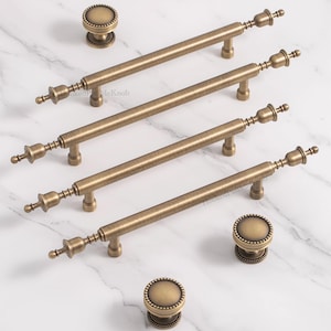 Può includere: Un set di ferramenta per mobili in ottone antico, composto da quattro maniglie lunghe a barra e tre pomelli rotondi. La ferramenta presenta un design dettagliato con estremità decorative e una finitura strutturata. I pezzi sono disposti su una superficie di marmo bianco. Le maniglie misurano circa 15 cm.