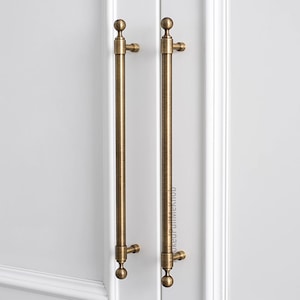 Puede incluir: Dos tiradores de armario de bronce antiguo con un diseño de barra vertical. Cada tirador tiene una bola en la parte superior e inferior, con un elemento decorativo en cada extremo. Los tiradores están montados en una puerta de armario blanca.