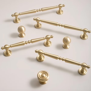 May include: A set of six gold-toned cabinet hardware pulls and knobs. The pulls are all different lengths and have a decorative design. The knobs are round and have a simple design.