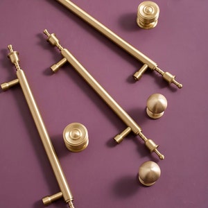 May include: Gold-toned cabinet hardware set, including long bar pulls, round knobs, and cylindrical knobs. The hardware has a brushed finish and is arranged on a purple surface. The set is suitable for kitchen or bathroom cabinets.