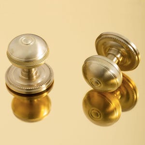 May include: Two brass door knobs with a round, textured design. The knobs have a circular base with a beaded edge and a smooth, reflective surface. The knobs are displayed on a gold surface.