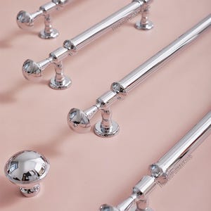 May include: Polished chrome cabinet hardware, including several bar pulls and a round knob. The pulls have a cylindrical design with rounded ends and a decorative base. The hardware is displayed on a light pink surface.