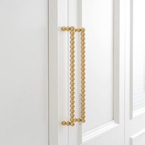 Könnte beinhalten: Ein goldfarbener Schrankgriff mit Perlendesign. Der Griff ist vertikal ausgerichtet und an einer weißen Schranktür befestigt. Der Griff hat eine rechteckige Form mit abgerundeten Ecken. Der Text "hookedoninteriordeb" ist sichtbar.