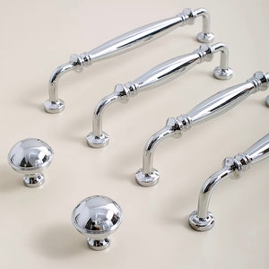 Puede incluir: Cuatro pomos de armario de cromo pulido y tres tiradores de armario de cromo. Los pomos son redondos y tienen una parte superior abovedada. Los tiradores son curvos y tienen un diseño decorativo.
