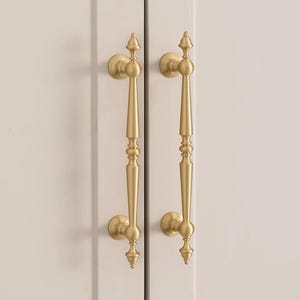 Może przedstawiać: Dwa złote uchwyty do szafek o klasycznym designie. Każdy uchwyt ma długi, smukły uchwyt z dekoracyjnymi detalami i zaokrągloną podstawą. Uchwyty są zamontowane na jasnych drzwiach szafki.