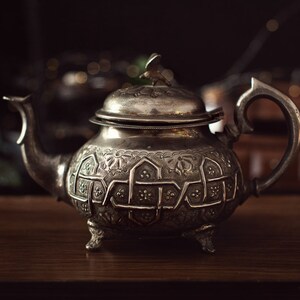 May include: Antique silver teapot with intricate embossed patterns. The teapot features a curved spout, a decorative handle, and a lid with a small bird-shaped finial. It rests on three small feet, showcasing a vintage design.