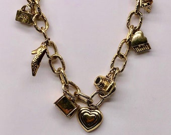 May include: Gold-toned chain necklace with various charms. Charms include a high-heeled shoe, a camera, a heart, a purse, and a cupcake. The chain has an oval link design and is displayed against a white background.
