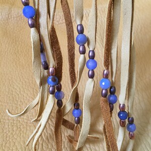May include: A close-up of a leather fringe with blue and brown beads. The fringe is made of light brown leather and the beads are strung on a cord.