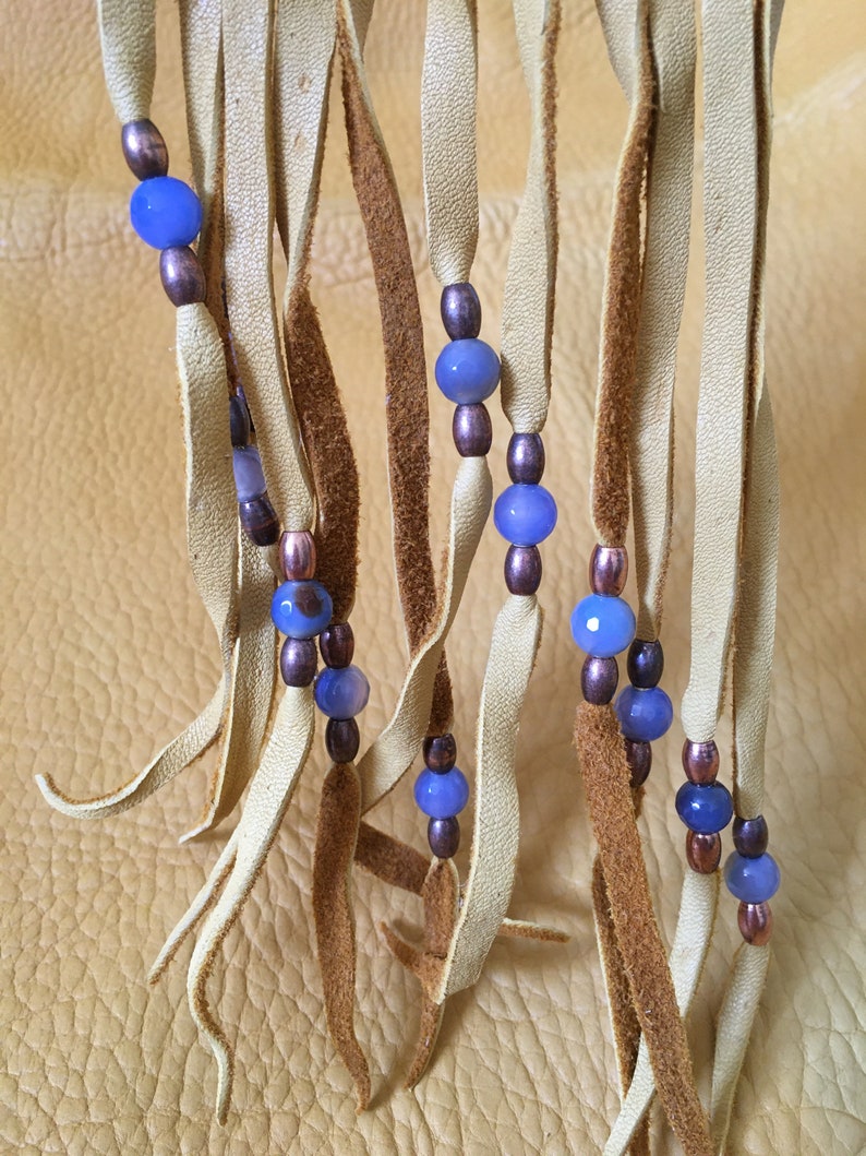 May include: A close-up of a leather fringe with blue and brown beads. The fringe is made of light brown leather and the beads are strung on a cord.