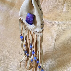 May include: A small, tan leather pouch with a purple and white stone attached. The pouch has a fringe of leather with blue and copper beads.