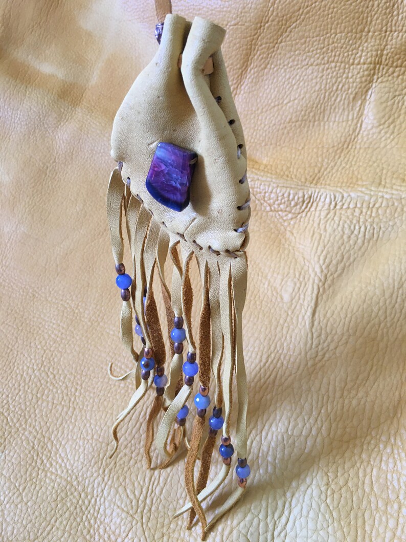 May include: A small, tan leather pouch with a purple and white stone attached. The pouch has a fringe of leather with blue and copper beads.