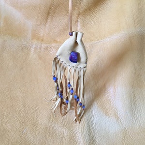 May include: A small white leather pouch with a blue and purple stone and fringe. The pouch is hanging from a string and is on a light brown leather background.