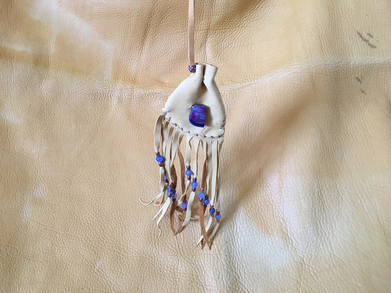 May include: A small white leather pouch with a blue and purple stone and fringe. The pouch is hanging from a string and is on a light brown leather background.